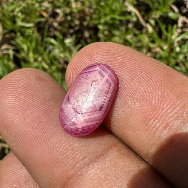 May include: An oval-shaped gemstone with a star-like pattern in pink and white. The gemstone has been cut and polished.