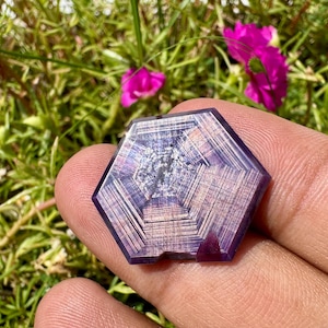 Könnte beinhalten: Ein sechseckiger Edelstein mit einer ausgeprägten violetten und rosafarbenen Farbpalette und einem sichtbaren geometrischen Muster aus konzentrischen Linien, die von der Mitte ausstrahlen. Der Stein hat eine glatte, polierte Oberfläche.