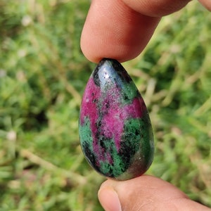 May include: A teardrop-shaped green, pink, and black gemstone pendant. The pendant is held between two fingers.