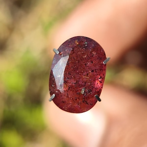 May include: A deep red oval gemstone with a faceted cut and inclusions. The gemstone is held in place by four prongs.