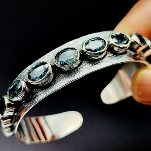 May include: A silver cuff bracelet with a textured finish and a row of oval-shaped blue gemstones.
