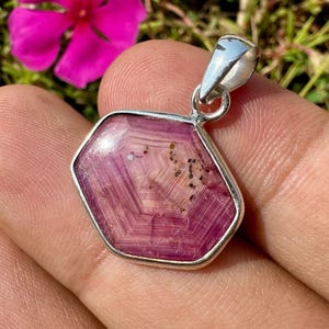 May include: A hexagonal pendant featuring a translucent pink gemstone with visible internal geometric banding and dark inclusions, set in a polished silver-toned metal frame with a small integrated bail at the top for attachment.