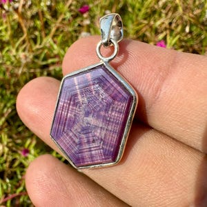 May include: A silver pendant with a large, faceted, purple gemstone. The gemstone has a unique, geometric pattern and is set in a silver bezel. The pendant is held up by a silver loop.