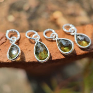 Op de afbeelding: Vier zilveren hangers in de vorm van een traan met groene edelsteenaccenten. De hangers liggen op een bruin oppervlak.