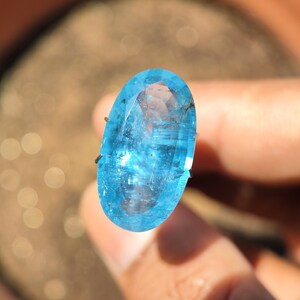 Puede incluir: Una piedra preciosa grande, de forma ovalada, de color azul claro con un corte facetado. La piedra preciosa se mantiene en su lugar mediante dos garras metálicas.