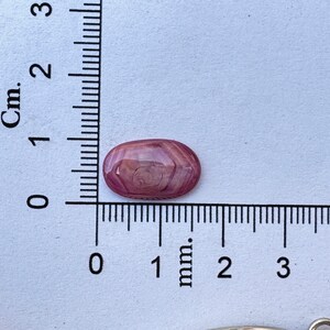 May include: A pink and white oval cabochon gemstone with a swirling pattern. The gemstone is approximately 1 cm long and is sitting on a white surface with a ruler.