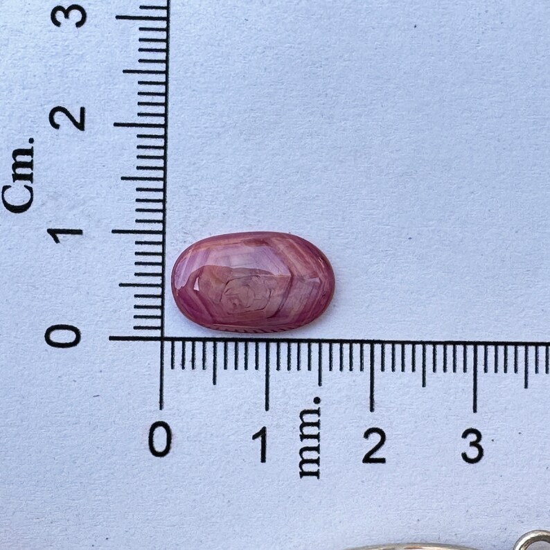 May include: A pink and white oval cabochon gemstone with a swirling pattern. The gemstone is approximately 1 cm long and is sitting on a white surface with a ruler.