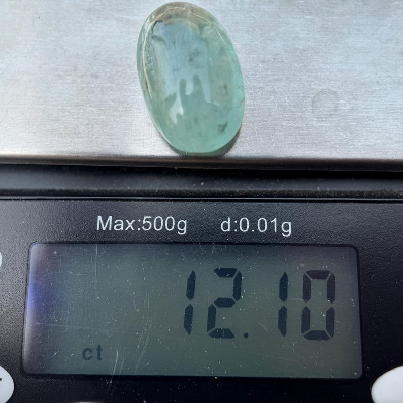 May include: A light blue oval gemstone weighing 12.10 carats on a digital scale.