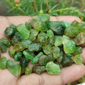 May include: A pile of green, rough, uncut gemstones. The stones are various sizes and shapes, with some having a slightly reddish hue.