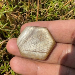 May include: A hexagonal, light brown gemstone with a star-like pattern of white lines. The gemstone is smooth and polished.