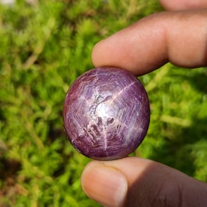May include: A close-up of a deep red star ruby cabochon with a six-pointed star pattern. The stone is smooth and polished, with a slight sheen.