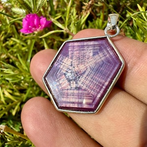 May include: A hexagonal pendant featuring a deep magenta and purple gemstone with a distinct geometric star-like pattern and visible internal striations, encased in a simple silver-toned metal frame with a small circular bail at the top.