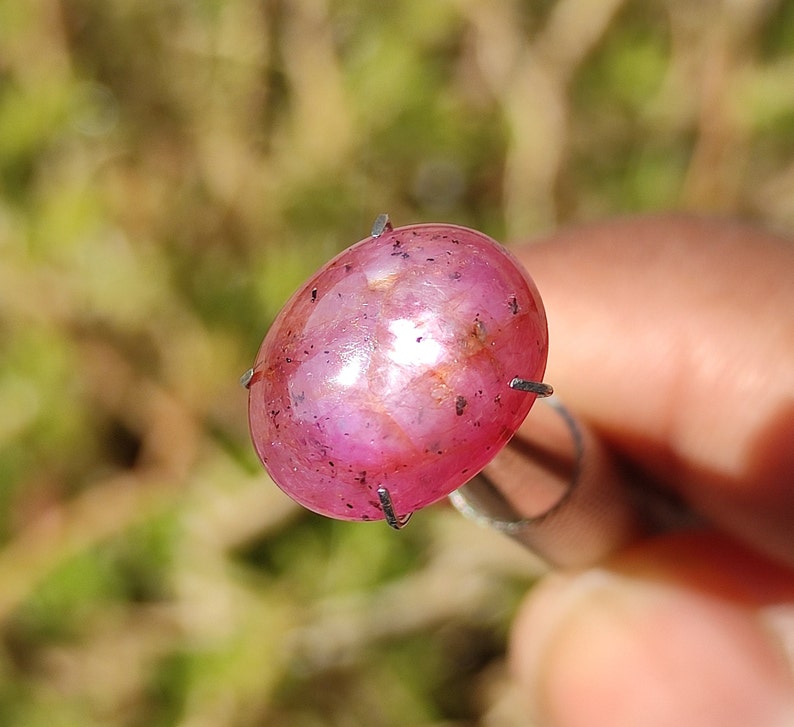 K&ouml;nnte beinhalten: Ein leuchtend rosa ovaler Edelstein mit einem sternf&ouml;rmigen Muster im Inneren. Der Edelstein ist in einer silbernen Krappenfassung gefasst.