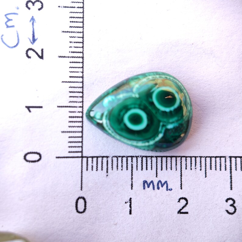 May include: A teardrop-shaped green gemstone with white and green swirls and two concentric circles in the center. The gemstone is approximately 1.5 cm long and is measured against a ruler.