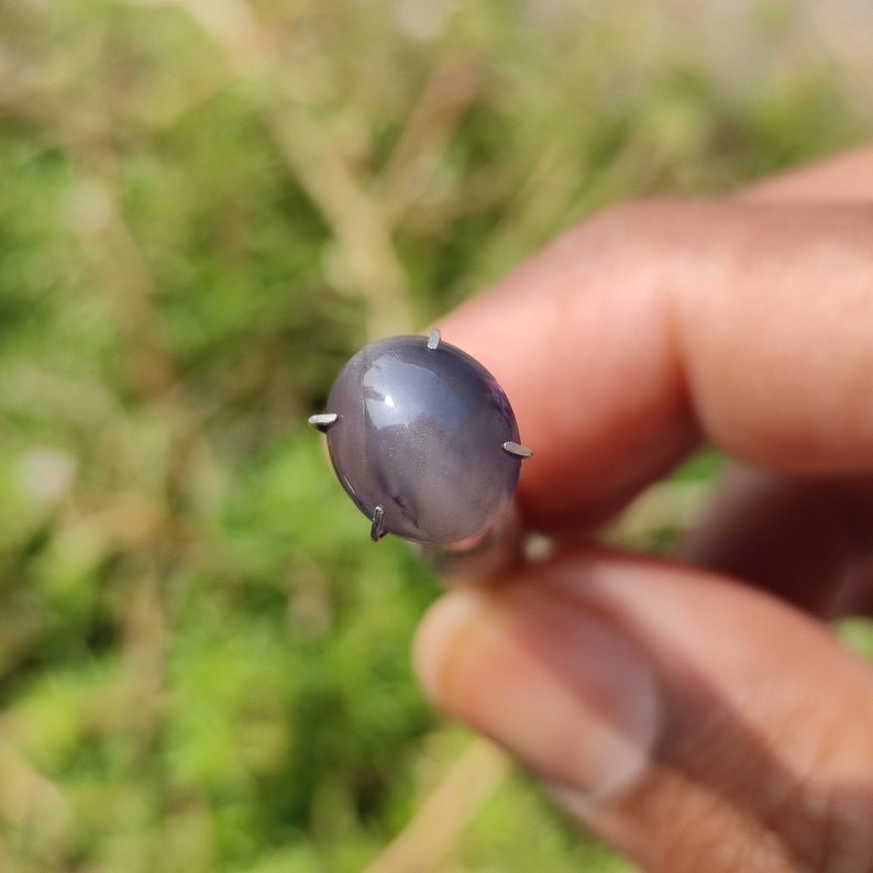 May include: A close-up of a gray oval-shaped gemstone with a metallic sheen. The gemstone is set in a prong setting with four prongs.