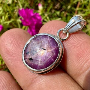 May include: A round, cabochon-cut star ruby pendant set in a polished silver bezel. The gemstone displays a deep magenta-purple hue with visible natural inclusions and a faint star effect, attached to a silver bail for necklace suspension.