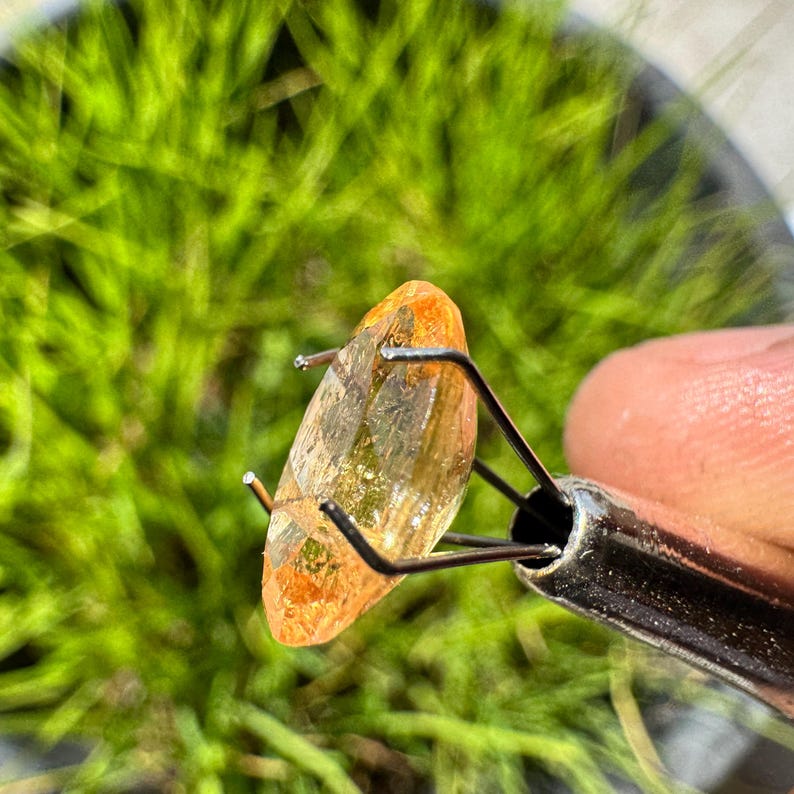 May include: A clear, faceted, orange gemstone held in a metal clamp. The gemstone has a slight yellow hue and is translucent.