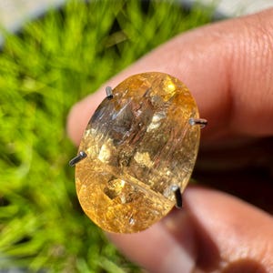 May include: A large, oval-shaped, golden citrine gemstone with a clear, glassy finish. The stone is set in a prong setting with four prongs.