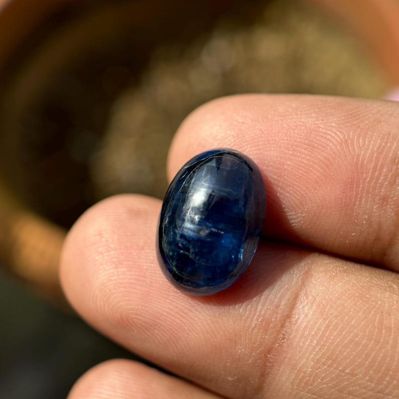 May include: A deep blue oval cabochon gemstone with a chatoyant effect. The stone is smooth and polished, with a slight shimmer.