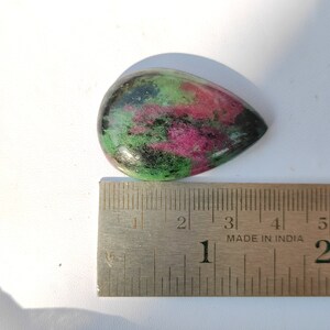 May include: A teardrop-shaped green, pink, and black gemstone cabochon measuring approximately 1 inch in length. The gemstone is placed next to a ruler for size reference.