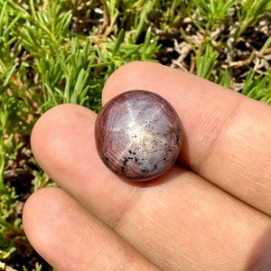 May include: A round, cabochon-cut, star ruby gemstone with a red and silver color. The stone has a star-like pattern in the center.