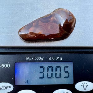 May include: A polished, irregular, dark brown agate stone with visible circular banding patterns. The stone is resting on a digital scale that displays a weight of 30.05 ct. The scale is black with a silver weighing platform and a blue backlit screen.