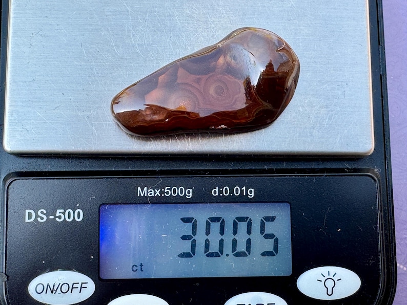 May include: A polished, irregular, dark brown agate stone with visible circular banding patterns. The stone is resting on a digital scale that displays a weight of 30.05 ct. The scale is black with a silver weighing platform and a blue backlit screen.