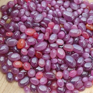 May include: A collection of polished, oval-shaped gemstones in varying shades of purple and red. The gemstones are scattered across a light brown wooden surface, with highlights and reflections visible on their smooth surfaces. The overall effect is a display of natural beauty and color.