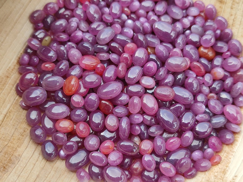 May include: A collection of polished, oval-shaped gemstones in varying shades of purple and red. The gemstones are scattered across a light brown wooden surface, with highlights and reflections visible on their smooth surfaces. The overall effect is a display of natural beauty and color.