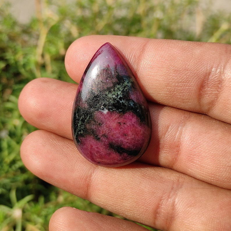 Puede incluir: Un colgante de piedra preciosa cabuj&oacute;n en forma de l&aacute;grima con un patr&oacute;n de color rosa y verde. La piedra est&aacute; pulida y tiene una superficie lisa.