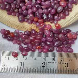 May include: A collection of polished, oval-shaped ruby gemstones in varying shades of red and pink. A metal ruler shows measurements in inches. The gemstones are scattered on a white surface and in a wooden container.
