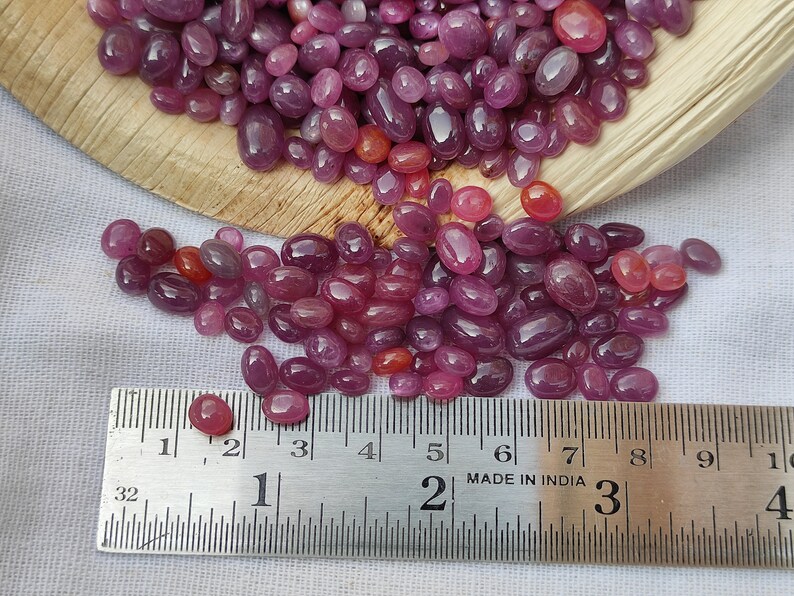 May include: A collection of polished, oval-shaped ruby gemstones in varying shades of red and pink. A metal ruler shows measurements in inches. The gemstones are scattered on a white surface and in a wooden container.
