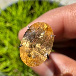 May include: A golden-yellow oval gemstone with a unique, striated pattern and flecks of gold. The gemstone is set in a prong setting with four prongs.