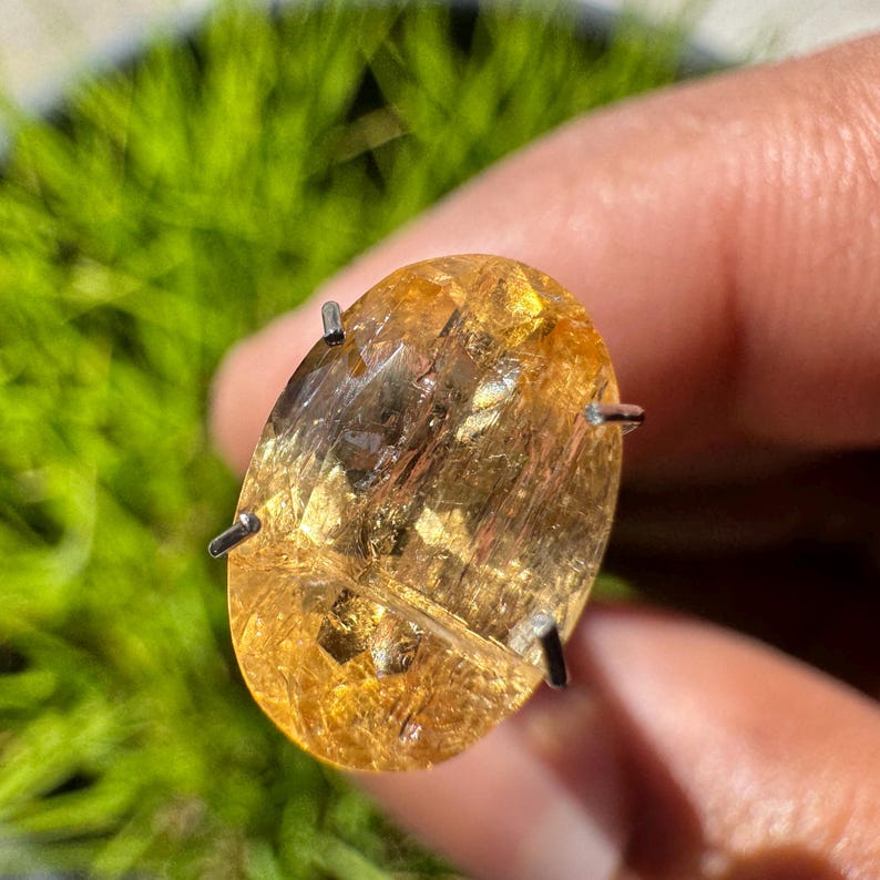 May include: A golden-yellow oval gemstone with a unique, striated pattern and flecks of gold. The gemstone is set in a prong setting with four prongs.