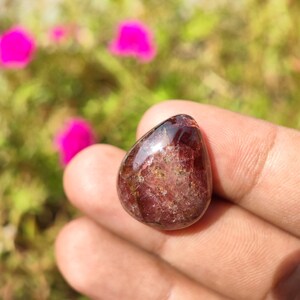 May include: A teardrop-shaped, red gemstone with a smooth, polished surface. The stone has a translucent appearance with visible inclusions.