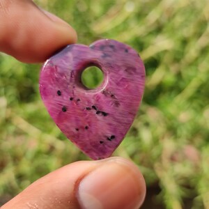 May include: A pink heart-shaped pendant with a hole in the center. The pendant is made of ruby and has black flecks throughout.