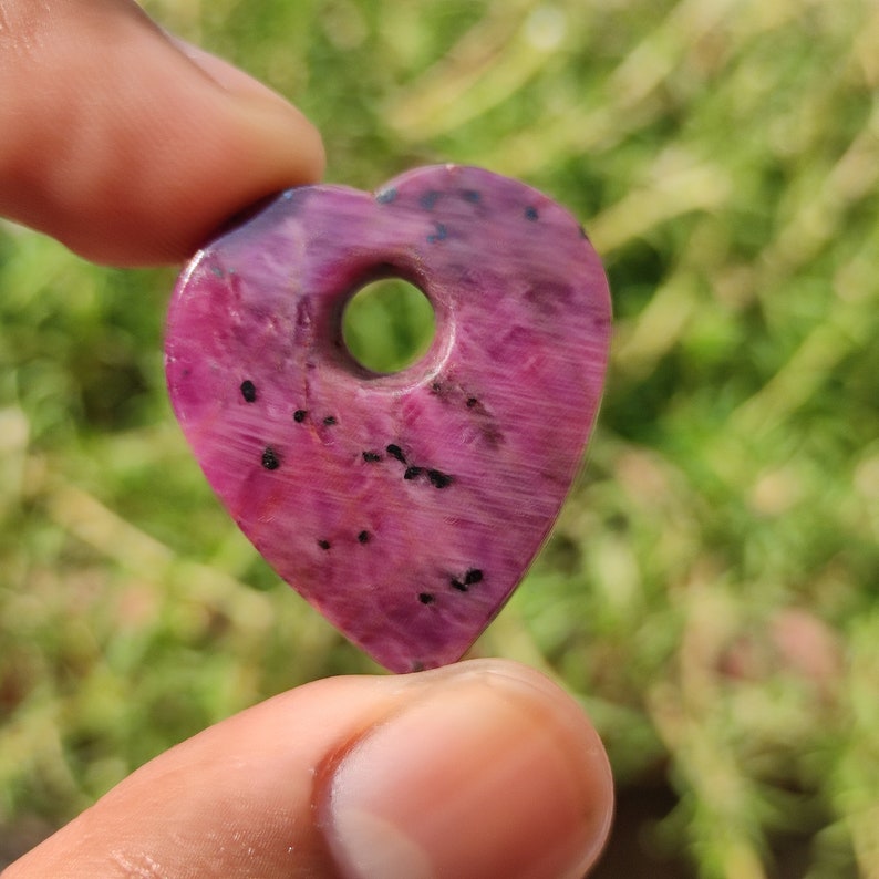 May include: A pink heart-shaped pendant with a hole in the center. The pendant is made of ruby and has black flecks throughout.