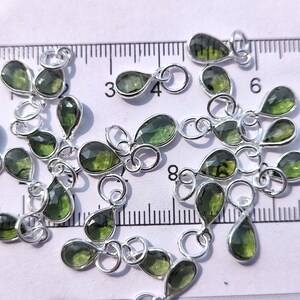 Op de afbeelding: Een groep van 20 groene edelsteen-traanbedels met zilveren springringen. De bedels zijn verspreid over een wit oppervlak met een liniaal op de achtergrond.