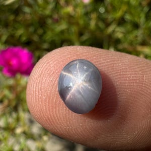 Puede incluir: Una piedra preciosa de zafiro estrellado gris con una estrella de seis puntas en el centro. La piedra preciosa tiene forma ovalada y una superficie lisa y pulida.