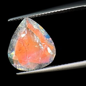 May include: A teardrop-shaped gemstone with a clear, faceted exterior and a pink, cloudy interior. The gemstone is held in a pair of tweezers.