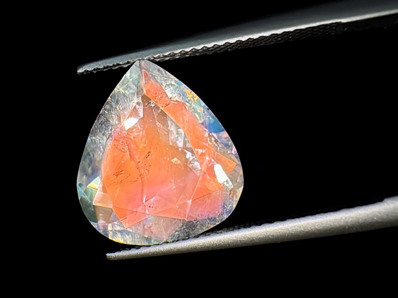 May include: A teardrop-shaped gemstone with a clear, faceted exterior and a pink, cloudy interior. The gemstone is held in a pair of tweezers.