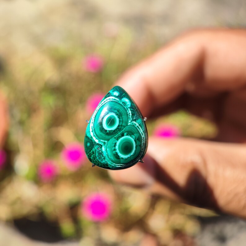 May include: A green and white malachite cabochon with a teardrop shape and intricate patterns. The stone is set in a silver bezel and is ready to be used in jewelry making.