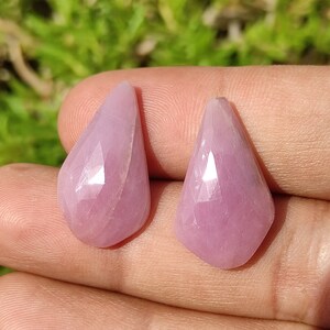 May include: Two faceted, teardrop-shaped gemstones in a soft pink hue. The gems have a polished, reflective surface, catching the light. The stones are held in a hand, with a blurred green background.