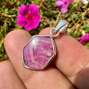 May include: A hexagonal pendant featuring a faceted, deep pink gemstone with visible internal banding patterns, set in a polished silver-toned metal frame with a simple circular bail at the top for attachment to a necklace chain.