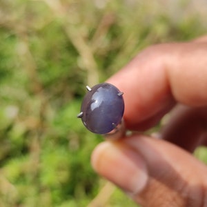 May include: A close-up of a gray oval-shaped gemstone with a star-like pattern in the center. The gemstone is set in a prong setting and is held by a hand.
