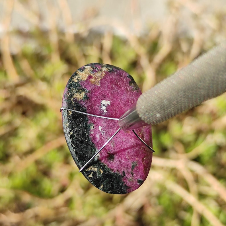 May include: An oval gemstone, predominantly magenta and black, secured with silver wire. The stone is being held by a silver tool. The background is blurred, suggesting an outdoor setting. The stone's surface appears polished.