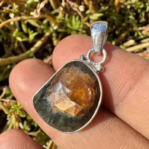 May include: A silver pendant featuring a large, faceted, dark green and brown gemstone. The gemstone has a unique, iridescent quality, with flashes of gold and green.