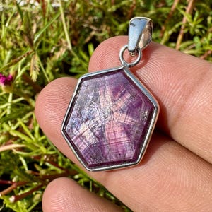 Op de afbeelding: Een zeshoekige hanger met een diep magenta sterrobijn edelsteen gezet in een gepolijst zilveren frame. De steen vertoont een duidelijk zesstralig sterpatroon over het gefacetteerde oppervlak, met een zilveren oogje aan de bovenzijde.