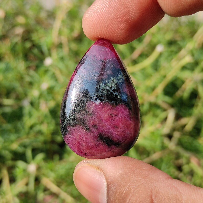 Puede incluir: Un colgante de piedra cabuj&oacute;n en forma de l&aacute;grima con un patr&oacute;n rosa y negro. La piedra tiene un acabado brillante y est&aacute; engastada en un engaste de tono plateado.