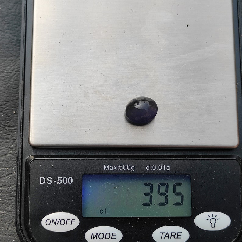 May include: A digital scale with a purple gemstone on the weighing platform. The scale reads "3.95 ct" and has the text "DS-500", "Max:500g", "d:0.01g", "ON/OFF", "MODE", and "TARE" on the display.